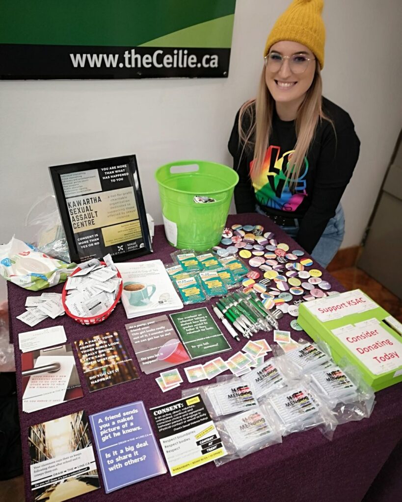 A staff sits next to a KSAC booth table overflowing with resources and swag items promoting sexual assault services