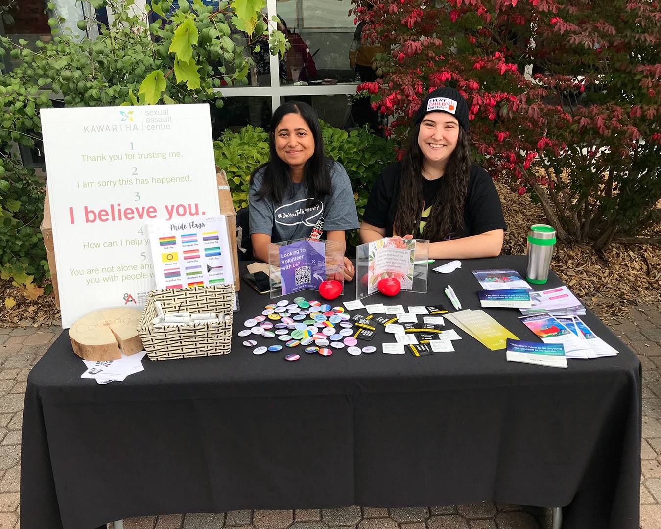 Two KSAC educators sit smiling at a KSAC booth filled with activities, buttons, and resources