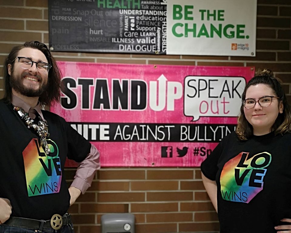 Two educators stand with hands on hips in t-shirts with the words "love wins" in rainbow motif, in front of a banner with the words "Stand Up Speak Out: Unite Against Bullying"