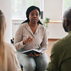 Young confident black woman with notebook and pen consulting intercultural couple sitting in front of her.