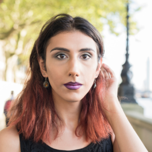 A person is staring directly at the viewer, you can see from their shoulders up. They are standing outside in front of a tree tucking hair behind one ear. They have dark shoulder-length hair with dyed pink ends, dark lipstick, and are in a black tank top.